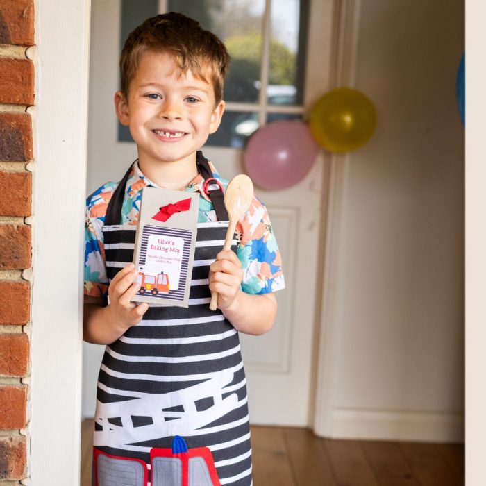 Boy wearing fire engine apron holding a wooden spoon and cookie mix gift box.